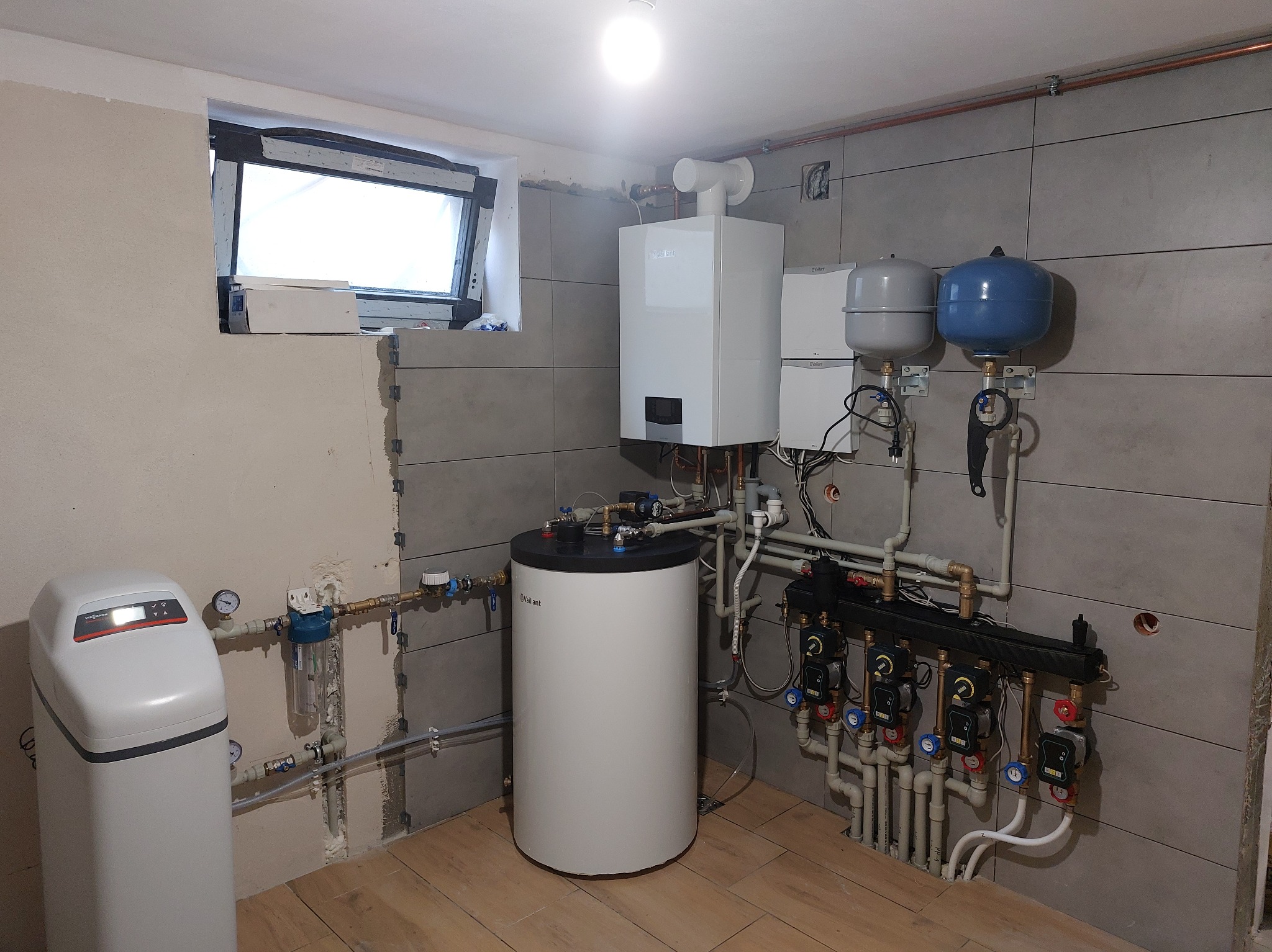 Nowoczesna kotłownia z piecem gazowym Vaillant, zbiornikiem, rozdzielaczem i zmiękczaczem wody. Szare płytki na ścianach, drewnopodobna podłoga, okno w tle.