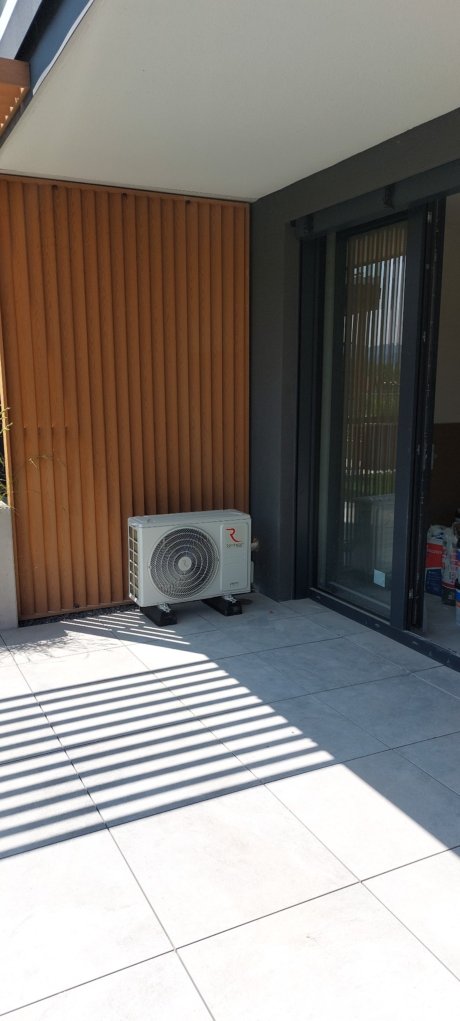 Biała jednostka zewnętrzna klimatyzacji Rotenso na balkonie z drewnianą ścianką i szarymi płytkami, oświetlona promieniami słońca padającymi przez żaluzje.