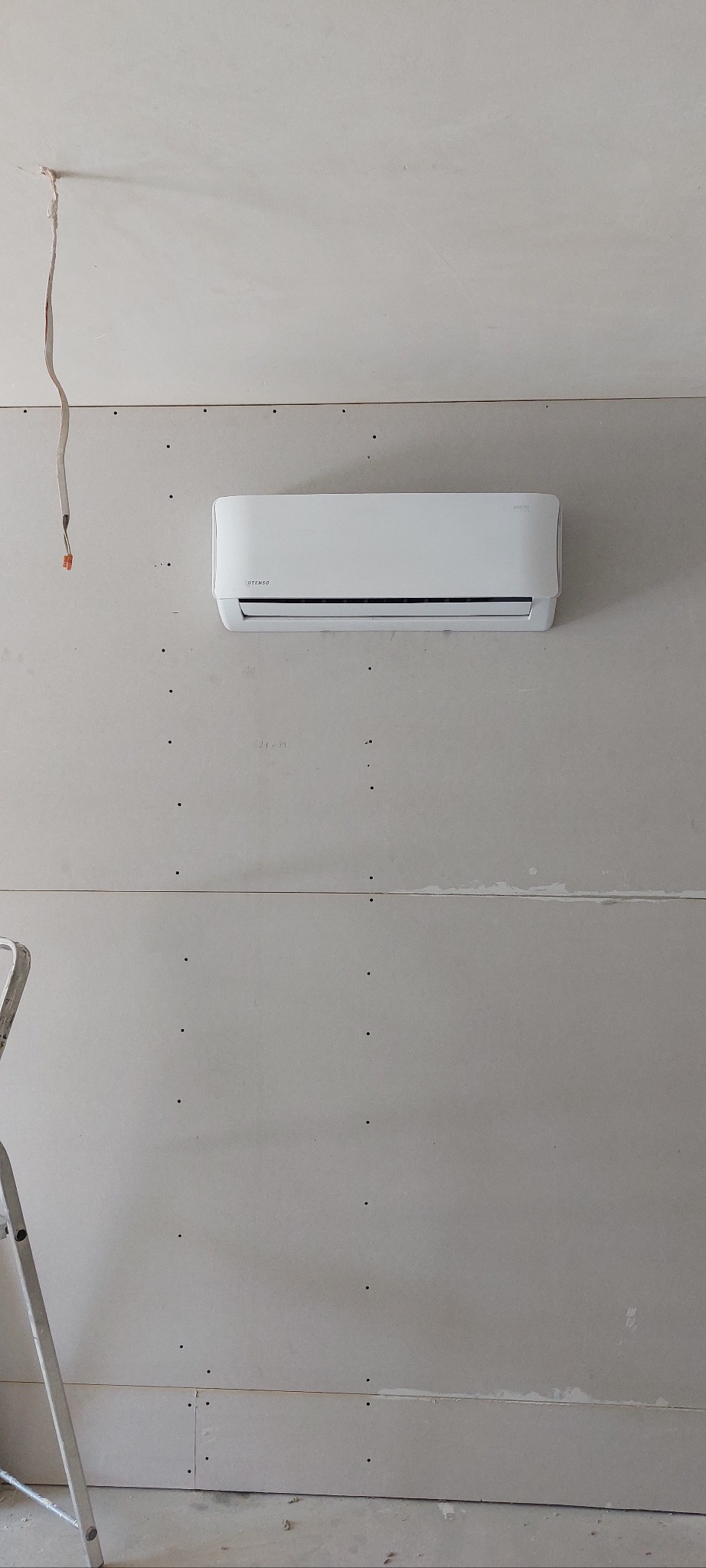 Biała klimatyzacja na ścianie z płyt kartonowo-gipsowych, widoczne ślady po montażu. Fragment drabiny malarskiej w lewym dolnym rogu. Kabel zwisający z sufitu.