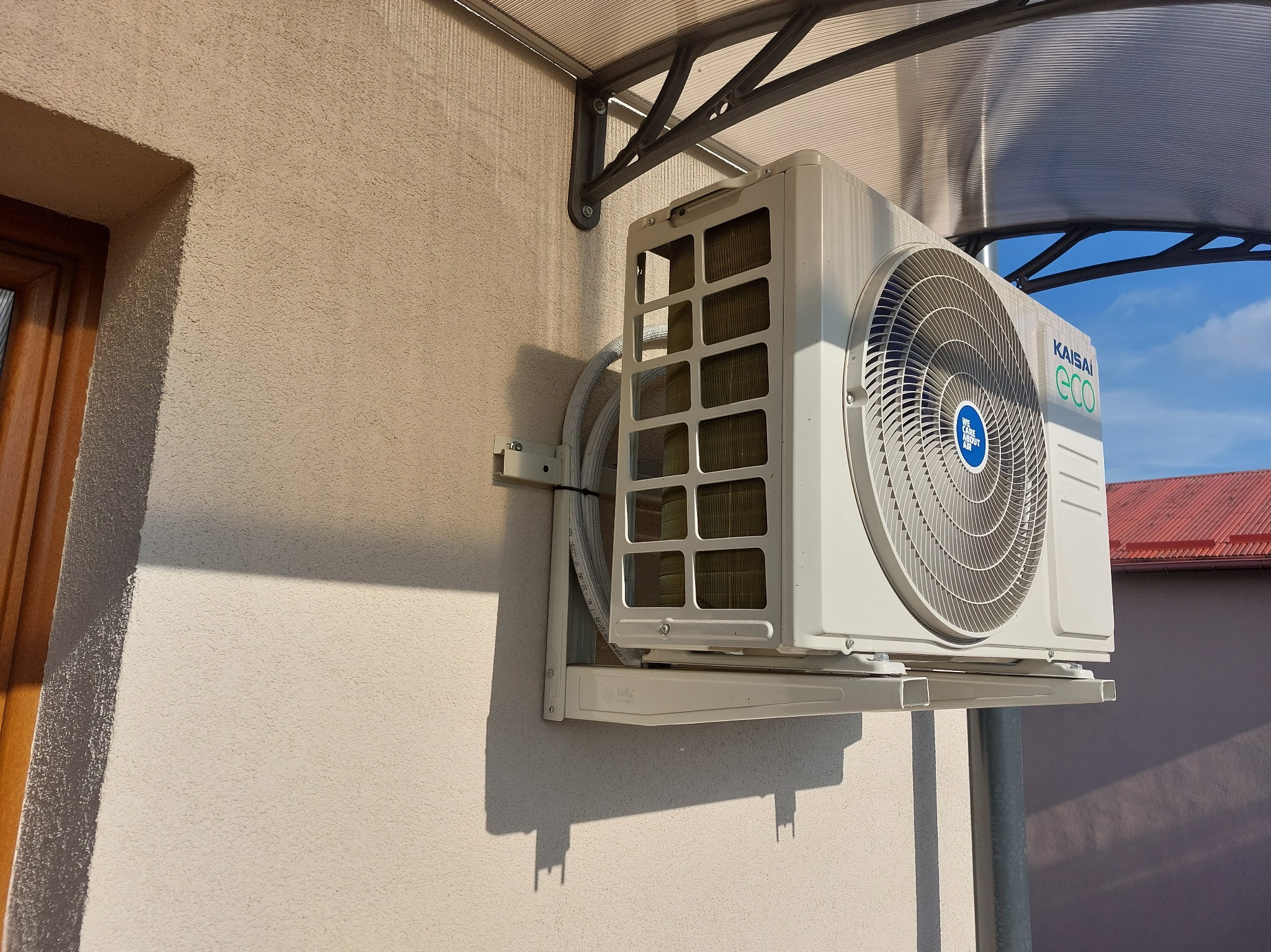Zamontowana jednostka zewnętrzna klimatyzacji Kaisai ECO na elewacji budynku, widoczna instalacja pod zadaszeniem. Słoneczny dzień.