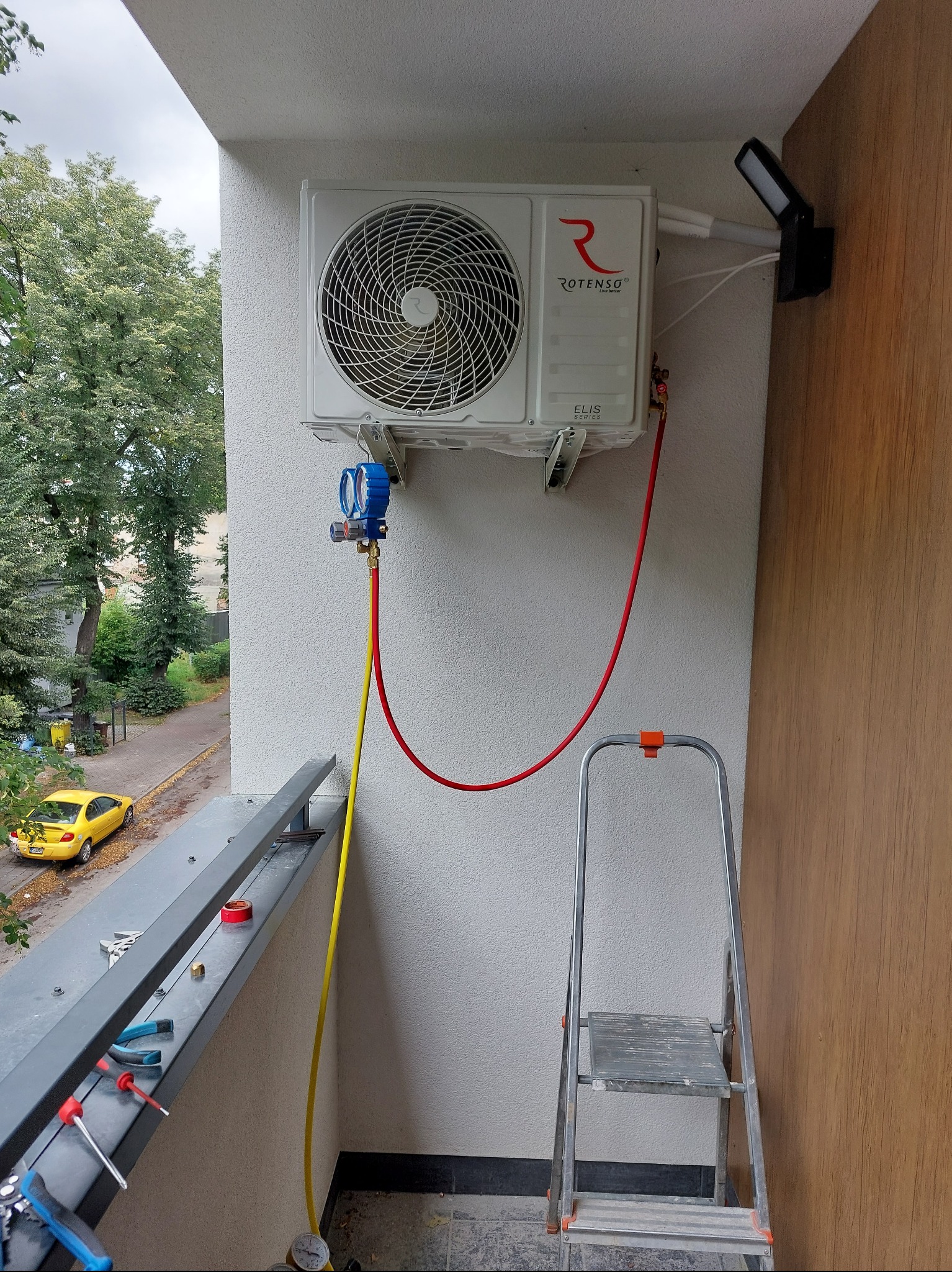 Biały klimatyzator Rotenso Elis Series zamontowany na balkonie, podłączony do manometrów i węży. Obok drabina, narzędzia na balustradzie. Widok na ulicę z żółtym autem.