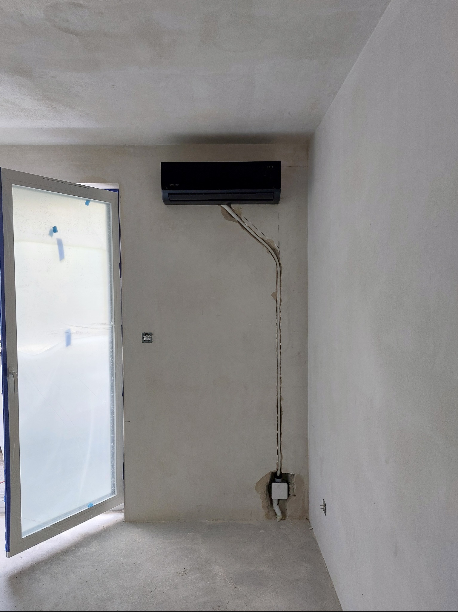 Czarny klimatyzator na ścianie w surowym wnętrzu. Widoczne przewody instalacyjne w bruzdach, otwarte drzwi balkonowe zabezpieczone folią malarską. Etap prac wykończeniowych.