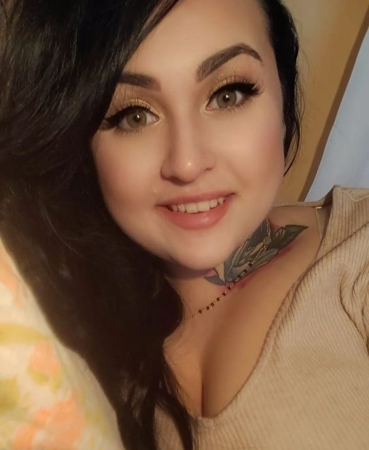 Uśmiechnięta brunetka z tatuażem na szyi, w beżowej bluzce z dekoltem w serek, z makijażem oczu w odcieniach złota i delikatnym naszyjnikiem.