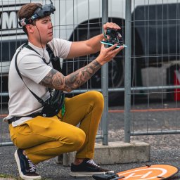 FlyDronRabikon Kacper Rabikowski - Mężczyzna w okularach FPV trzyma drona wyścigowego, przygotowując się do lotu. W tle siatka i biały bus. Widoczny lądowisko dla drona.