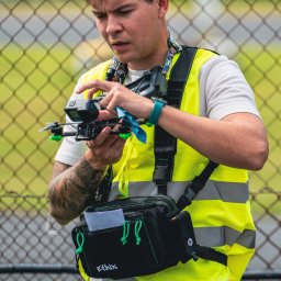 FlyDronRabikon Kacper Rabikowski - Młody mężczyzna w kamizelce ostrzegawczej przygotowuje drona z kamerą GoPro przed lotem. W tle siatka ogrodzeniowa i zieleń.