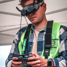 FlyDronRabikon Kacper Rabikowski - Operator drona FPV w kamizelce ostrzegawczej, z goglami i kontrolerem w dłoniach, gotowy do lotu. Widoczne logo Wallrav Race Center.