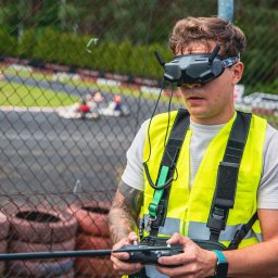 FlyDronRabikon Kacper Rabikowski - Operator drona FPV w kamizelce ostrzegawczej, sterujący dronem na torze kartingowym. W tle gokarty i opony. Ujęcie z bliska, skupione na operatorze i jego kontrolerze.