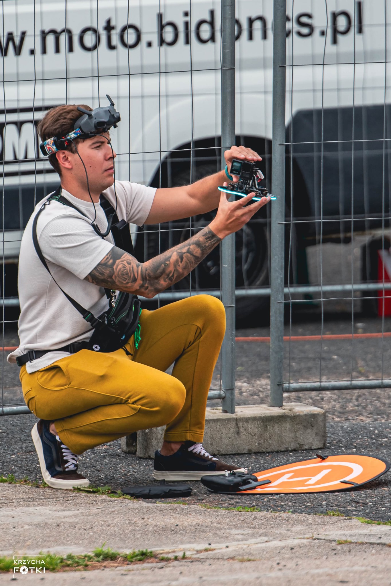 Mężczyzna w okularach FPV trzyma drona wyścigowego, przygotowując się do lotu. W tle siatka i biały bus. Widoczny lądowisko dla drona.