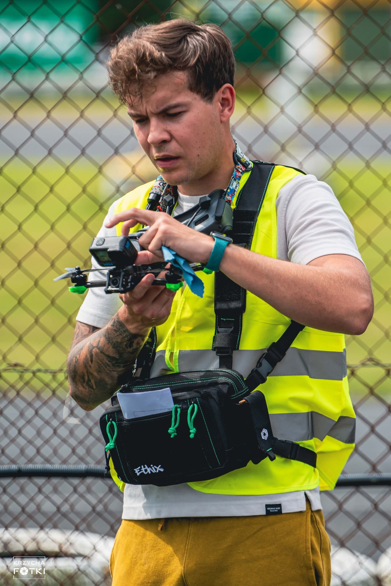 Młody mężczyzna w kamizelce ostrzegawczej przygotowuje drona z kamerą GoPro przed lotem. W tle siatka ogrodzeniowa i zieleń.