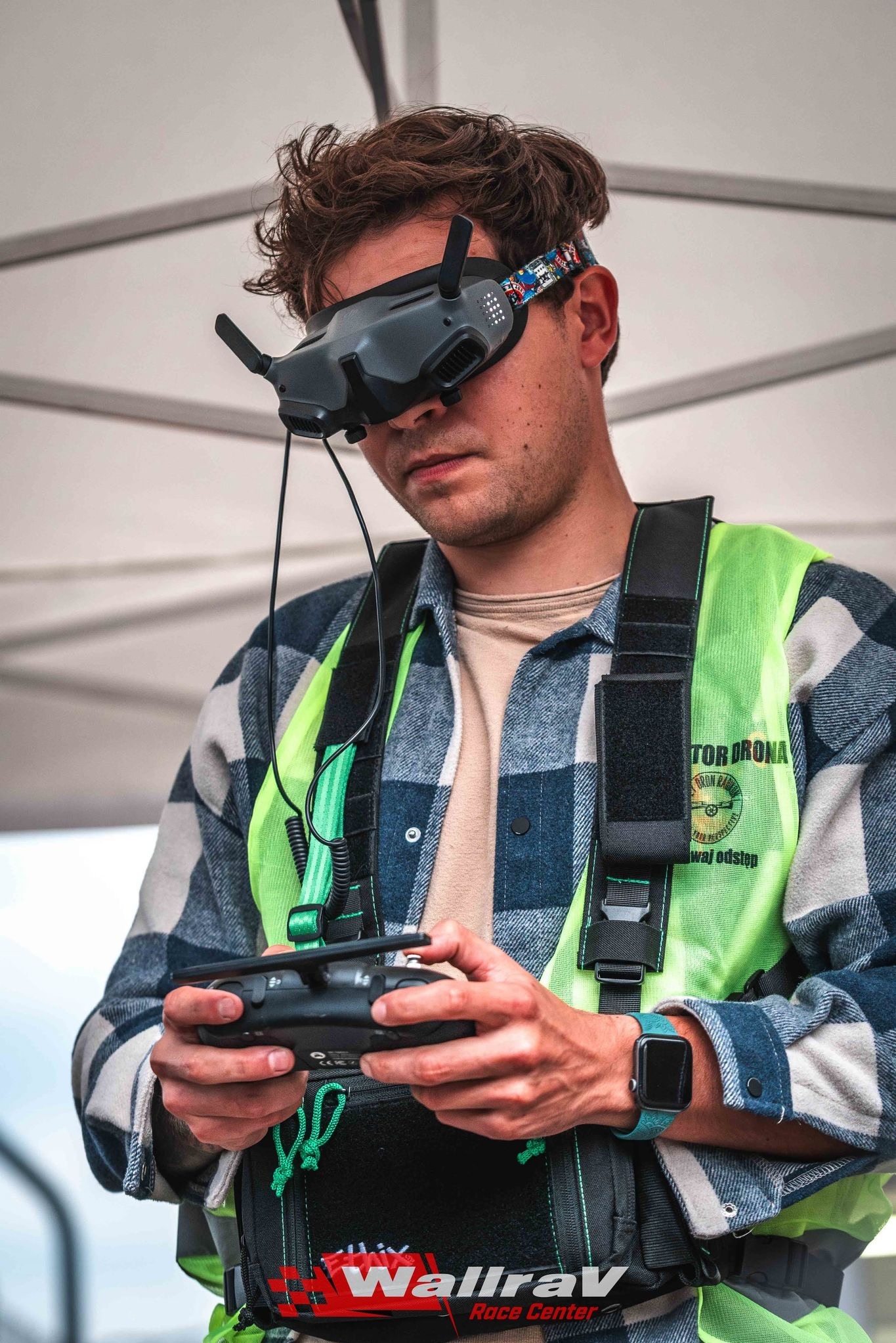 Operator drona FPV w kamizelce ostrzegawczej, z goglami i kontrolerem w dłoniach, gotowy do lotu. Widoczne logo Wallrav Race Center.