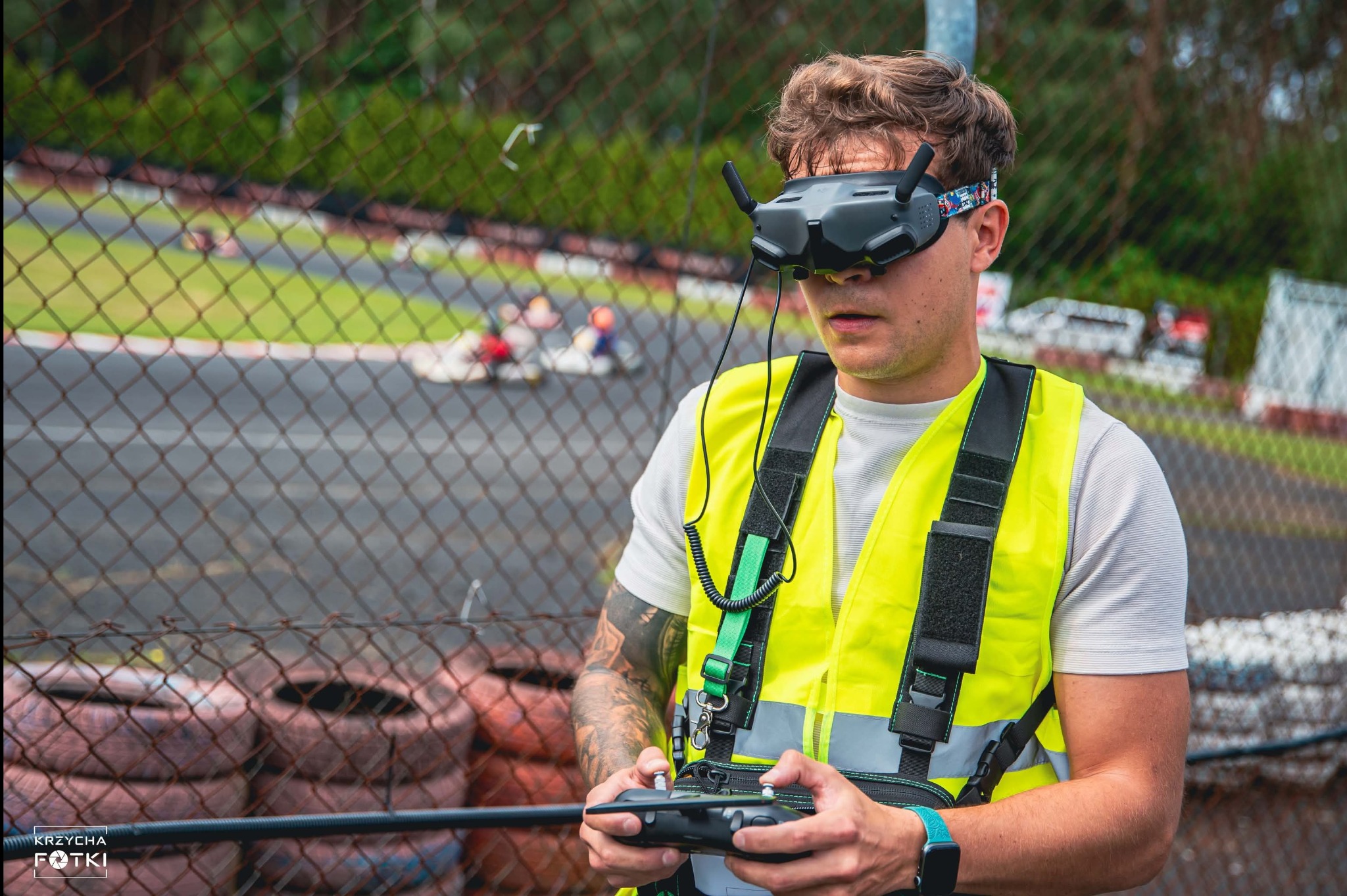 Operator drona FPV w kamizelce ostrzegawczej, sterujący dronem na torze kartingowym. W tle gokarty i opony. Ujęcie z bliska, skupione na operatorze i jego kontrolerze.