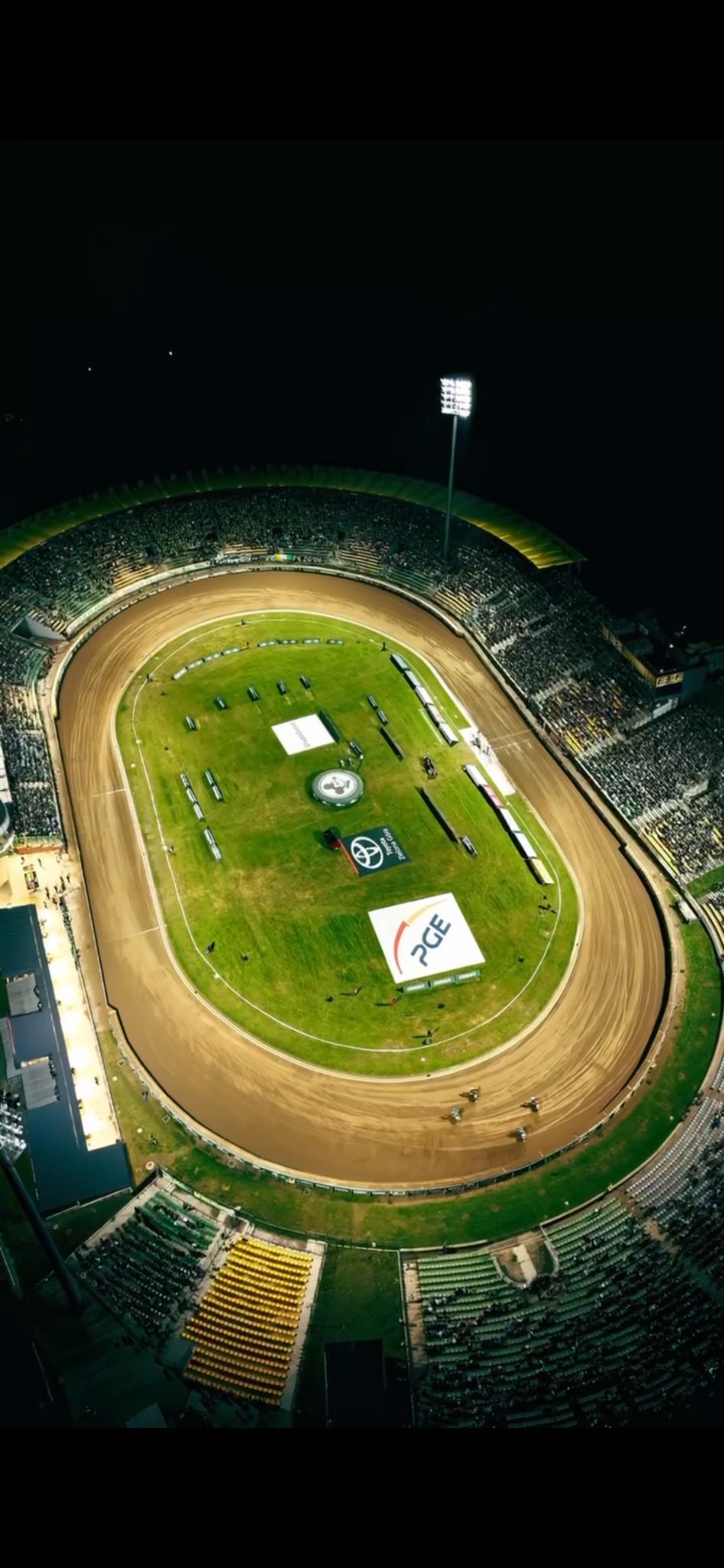 Nocny widok z drona na stadion żużlowy podczas zawodów, oświetlony reflektorami, z widocznymi logotypami sponsorów na murawie i trybunami wypełnionymi kibicami.