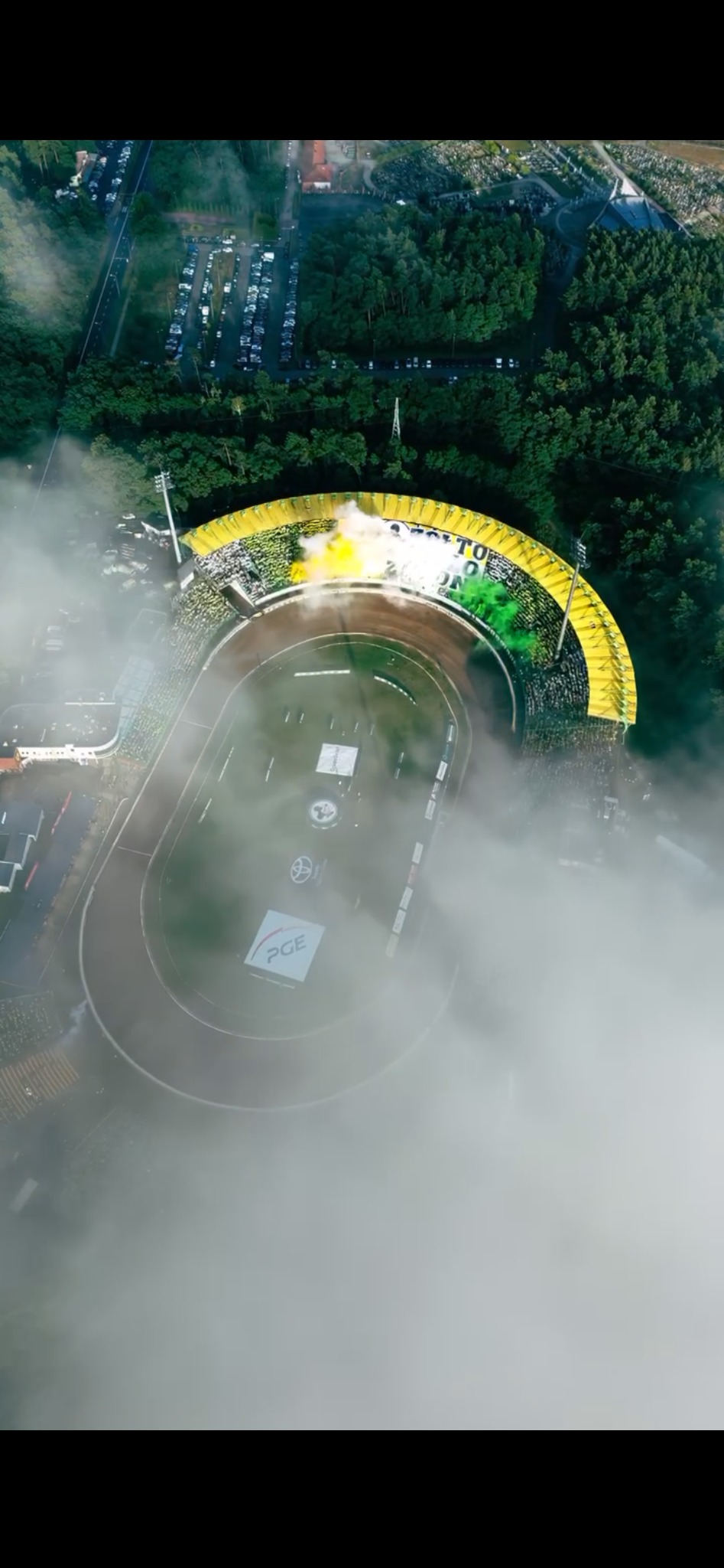 Widok z drona na stadion żużlowy podczas wydarzenia sportowego, otoczony zielenią i częściowo przesłonięty mgłą. Kibice na trybunach tworzą kolorową oprawę.