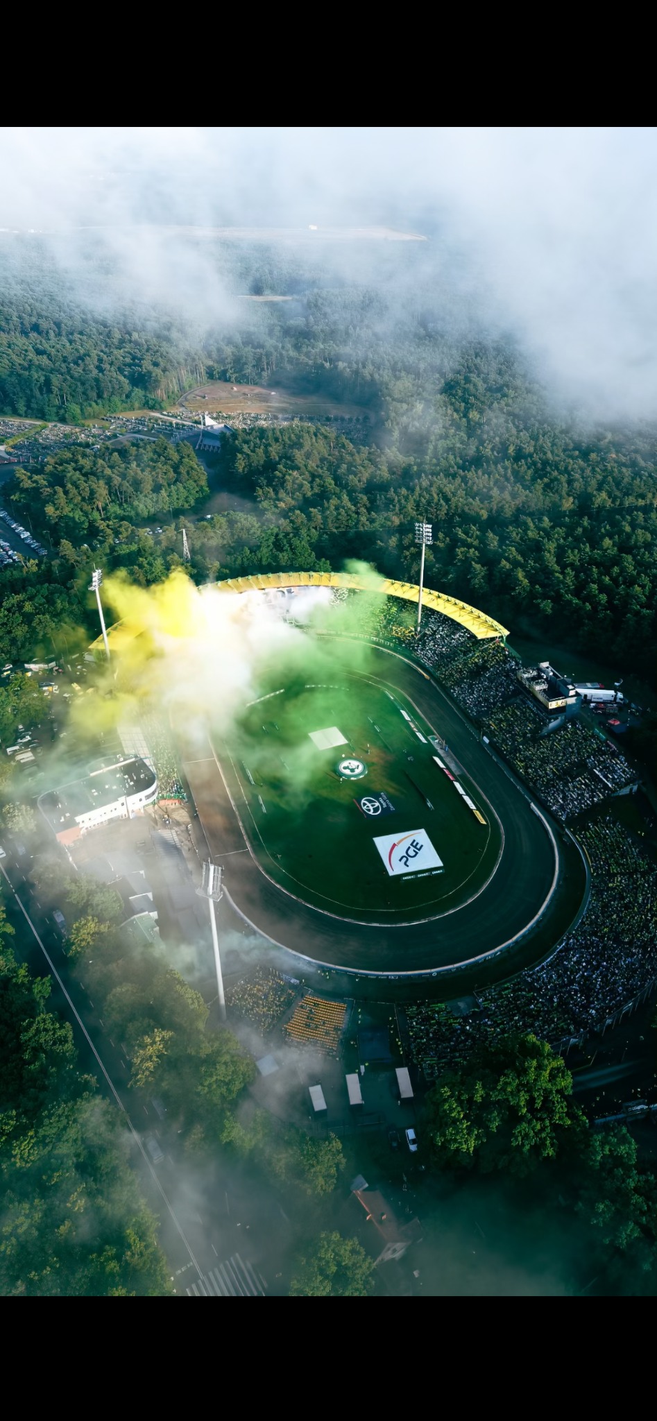 Widok z drona na stadion żużlowy podczas zawodów, otoczony lasem i mgłą, z widocznymi kibicami i efektami pirotechnicznymi w kolorach żółtym i zielonym.