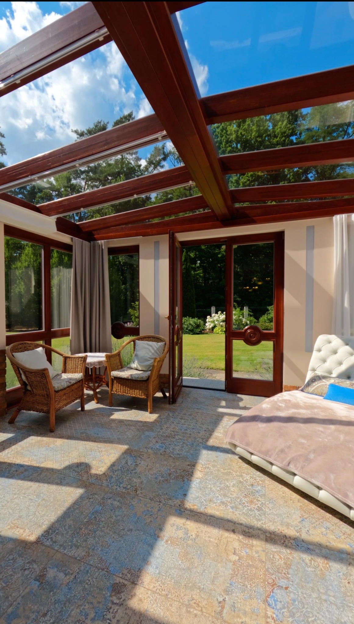 Jasna oranżeria z drewnianą konstrukcją i widokiem na ogród. Wnętrze z wiklinowymi fotelami, stolikiem i leżanką. Dekoracyjna podłoga w odcieniach beżu i błękitu.