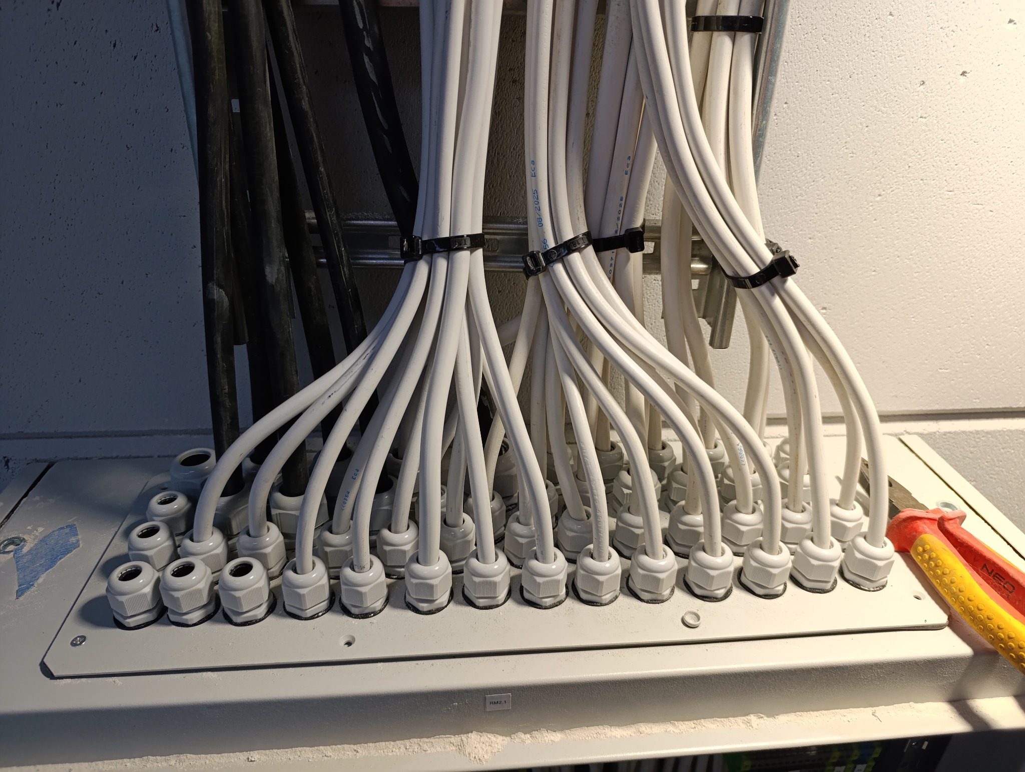 Uporządkowane białe przewody elektryczne wchodzące do skrzynki rozdzielczej, zabezpieczone czarnymi opaskami zaciskowymi. Widoczne narzędzia.