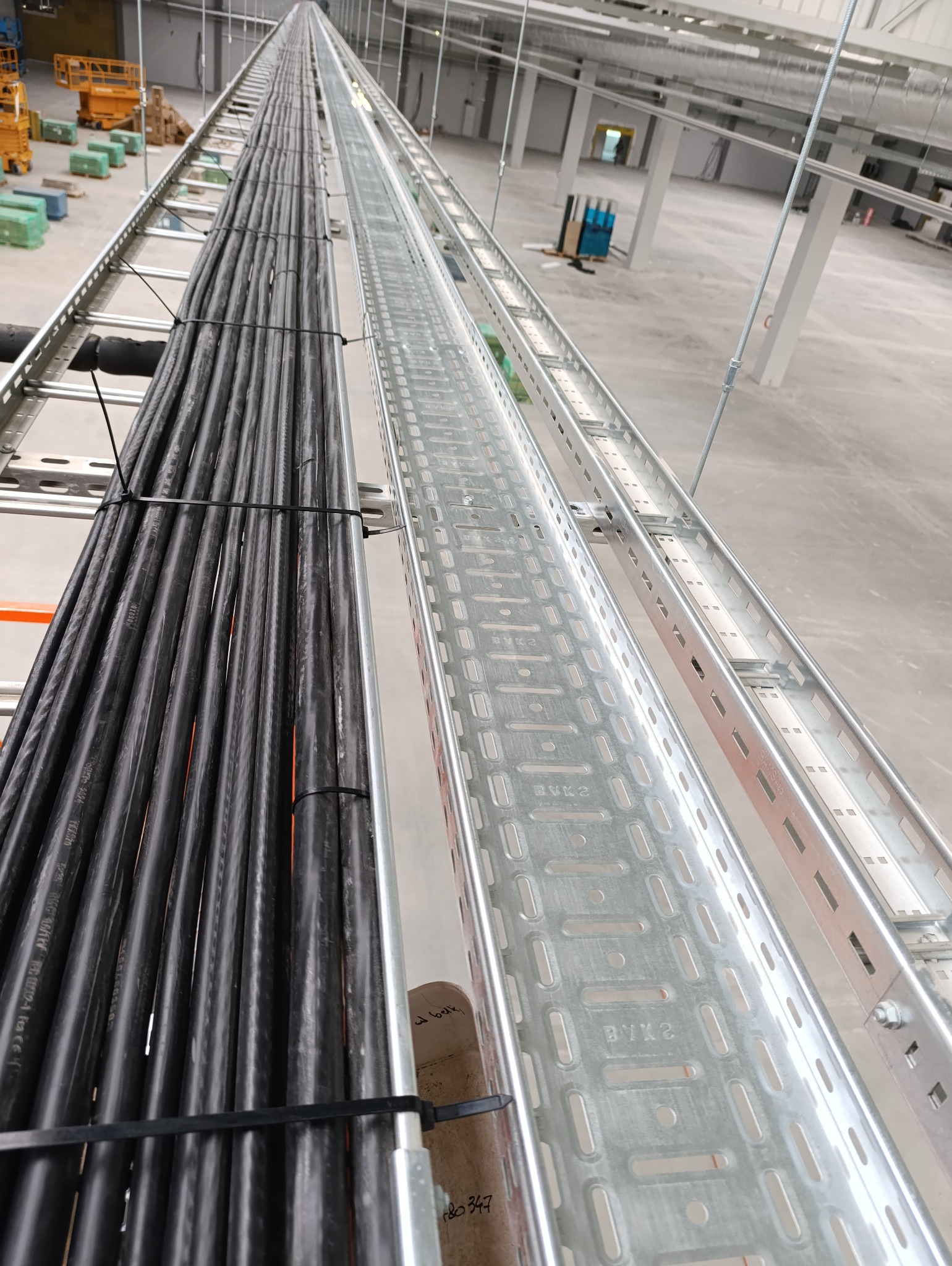 Długi ciąg czarnych kabli ułożonych w metalowej korycie instalacyjnej, widok z góry w hali przemysłowej. Kable spięte opaskami.