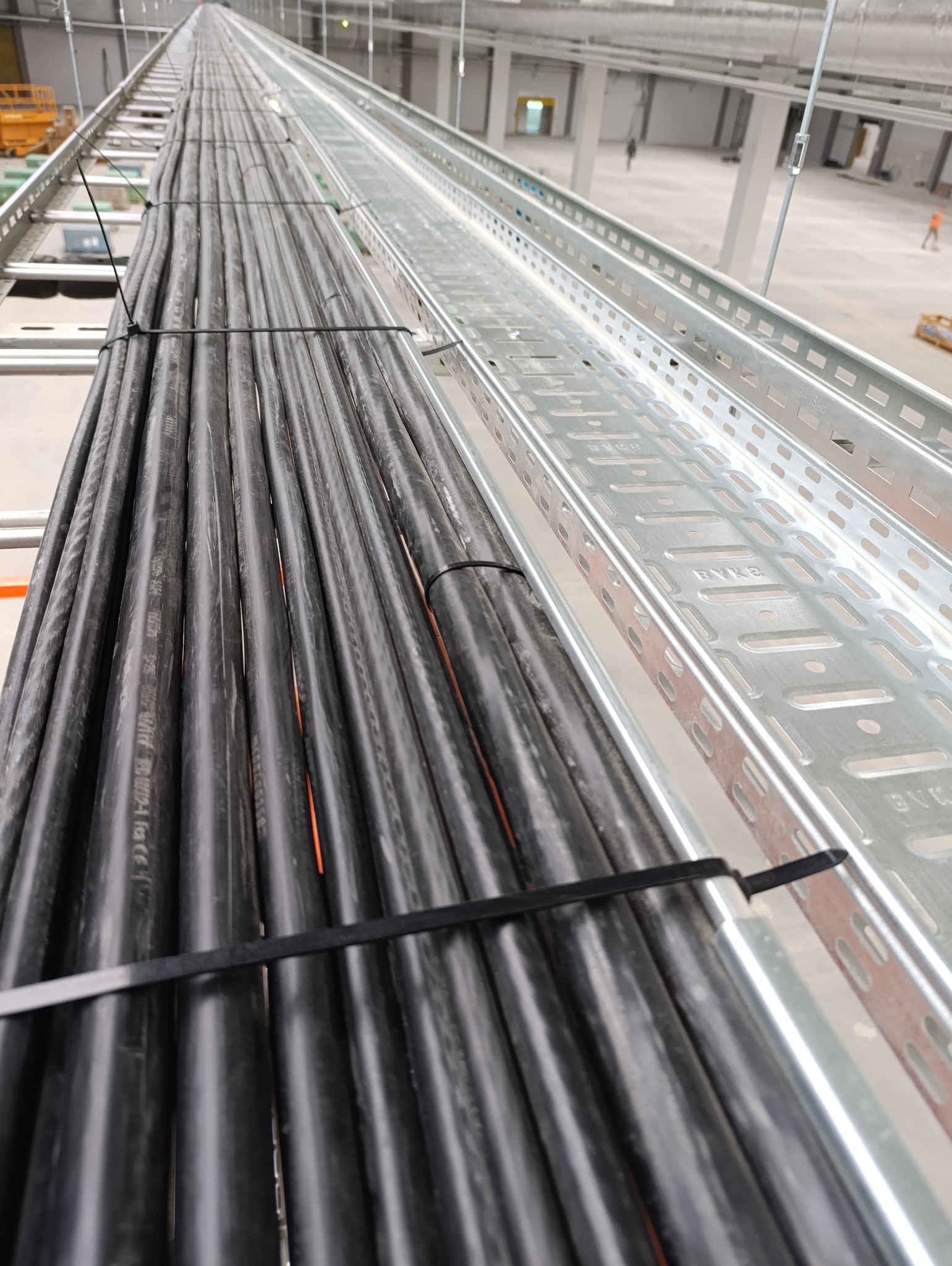 Ułożone kable elektryczne w korytkach kablowych, zabezpieczone opaskami. Widok z góry, perspektywa zbieżna, hala przemysłowa w tle.