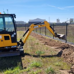PHU Maciej Kaźmierczak - Żółta mini koparka JCB wykonuje wykop wzdłuż ogrodzenia panelowego na trawiastym terenie. Widoczne budynki w tle i błękitne niebo.