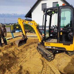 PHU Maciej Kaźmierczak - Żółta minikoparka JCB 16C-1 w trakcie prac ziemnych na piaszczystym terenie. Operator wyrównuje teren łopatą. W tle widoczny budynek mieszkalny i ogrodzenie.