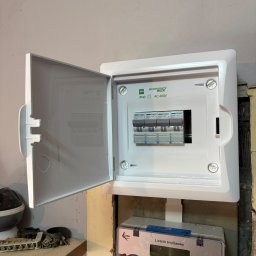MD - SYSTEM Mateusz Drabik - Otwarta skrzynka elektryczna z bezpiecznikami i licznikiem trójfazowym, zamontowana na ścianie. Widoczne detale instalacji i oznaczenia na urządzeniach.