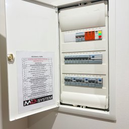 MD - SYSTEM Mateusz Drabik - Otwarta, biała skrzynka rozdzielcza Hager z bezpiecznikami i schematem obwodów na ścianie. Widoczne kontrolki faz, wyłączniki różnicowoprądowe i logo MD-System.