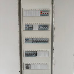 MD - SYSTEM Mateusz Drabik - Rozdzielnica elektryczna Hager w surowej wnęce ściany, widoczne zabezpieczenia, gniazdo. Część obwodów włączona, sygnalizowana czerwonymi diodami. Instalacja w trakcie prac wykończeniowych.