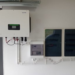 MD - SYSTEM Mateusz Drabik - Biały inwerter SOFAR i rozdzielnice elektryczne zamontowane na ścianie, połączone białymi rurkami instalacyjnymi. Widok z przodu, oświetlenie dzienne.