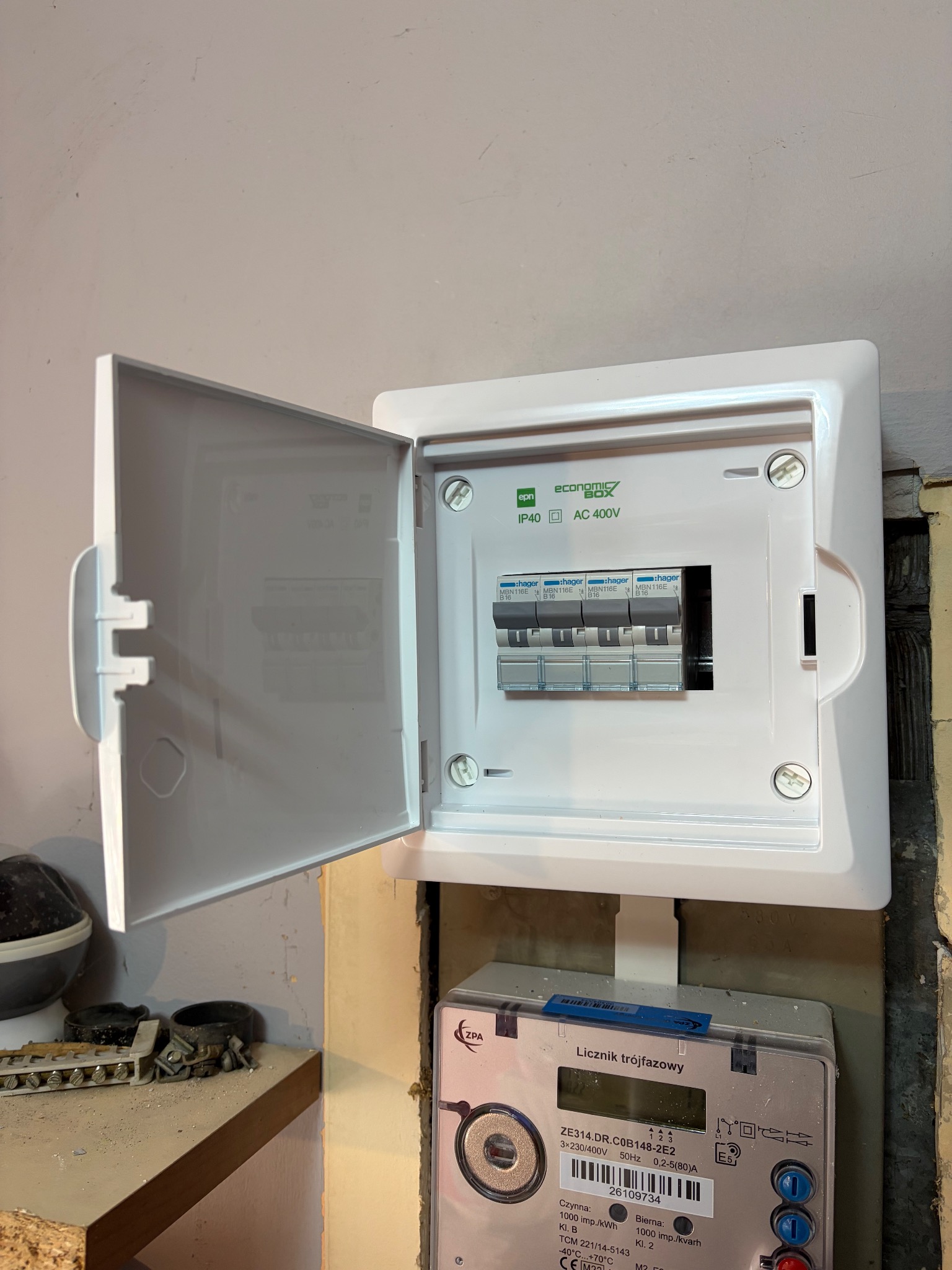 Otwarta skrzynka elektryczna z bezpiecznikami i licznikiem trójfazowym, zamontowana na ścianie. Widoczne detale instalacji i oznaczenia na urządzeniach.