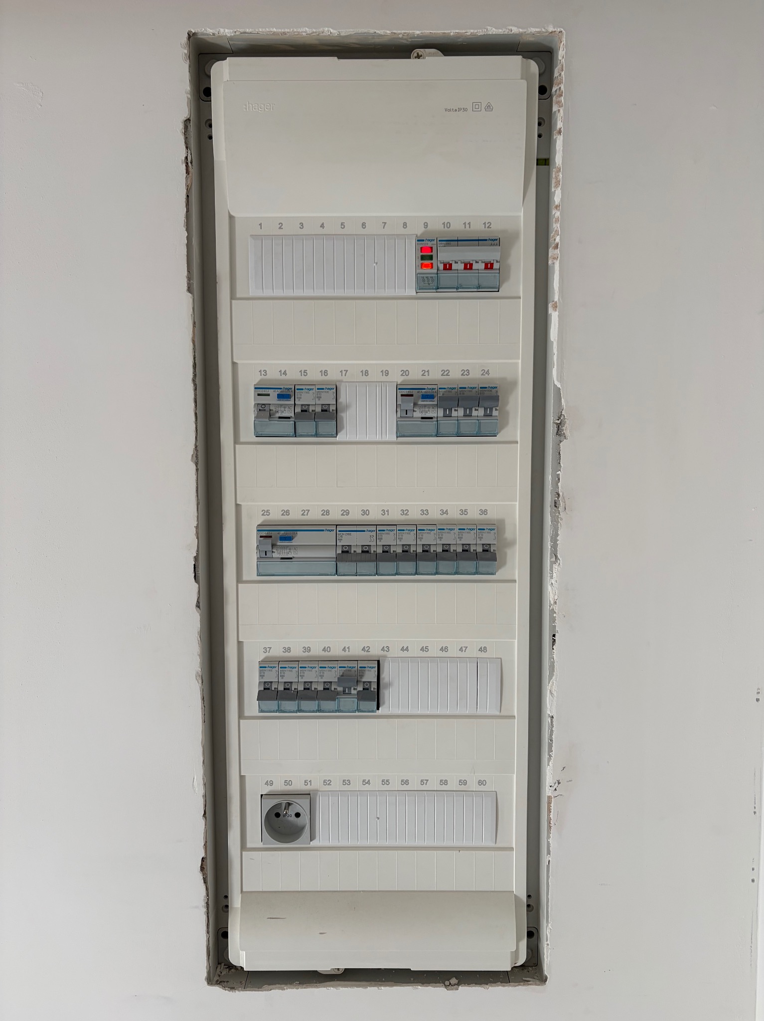 Rozdzielnica elektryczna Hager w surowej wnęce ściany, widoczne zabezpieczenia, gniazdo. Część obwodów włączona, sygnalizowana czerwonymi diodami. Instalacja w trakcie prac wykończeniowych.