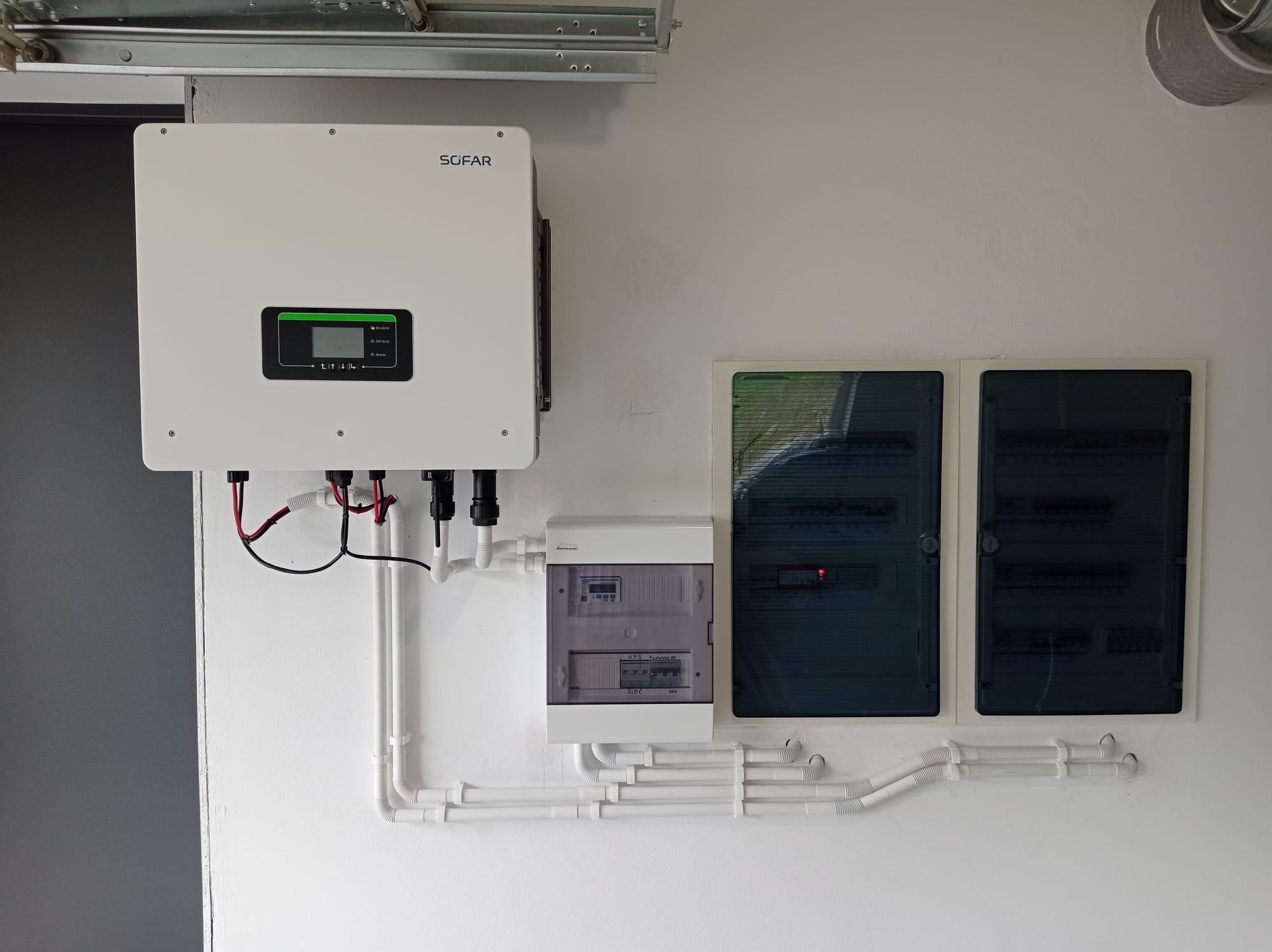 Biały inwerter SOFAR i rozdzielnice elektryczne zamontowane na ścianie, połączone białymi rurkami instalacyjnymi. Widok z przodu, oświetlenie dzienne.