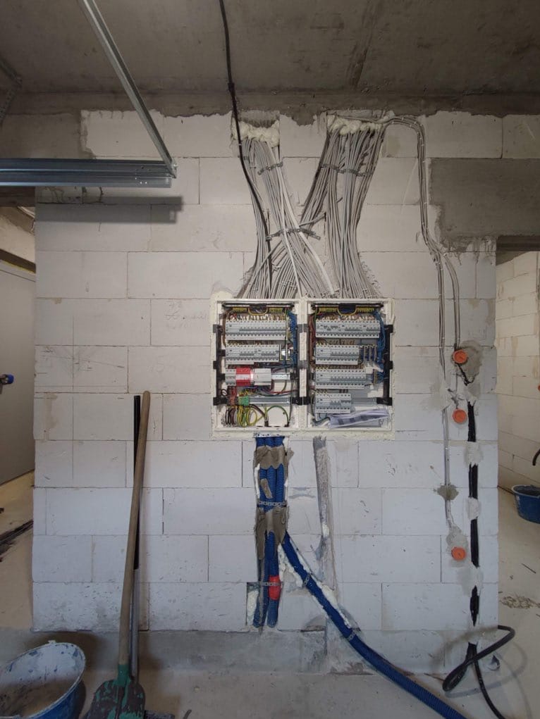 Rozdzielnia elektryczna z licznymi przewodami w surowym wnętrzu. Widoczne kable w peszlach, puszki instalacyjne i narzędzia. Ściany z betonu komórkowego.