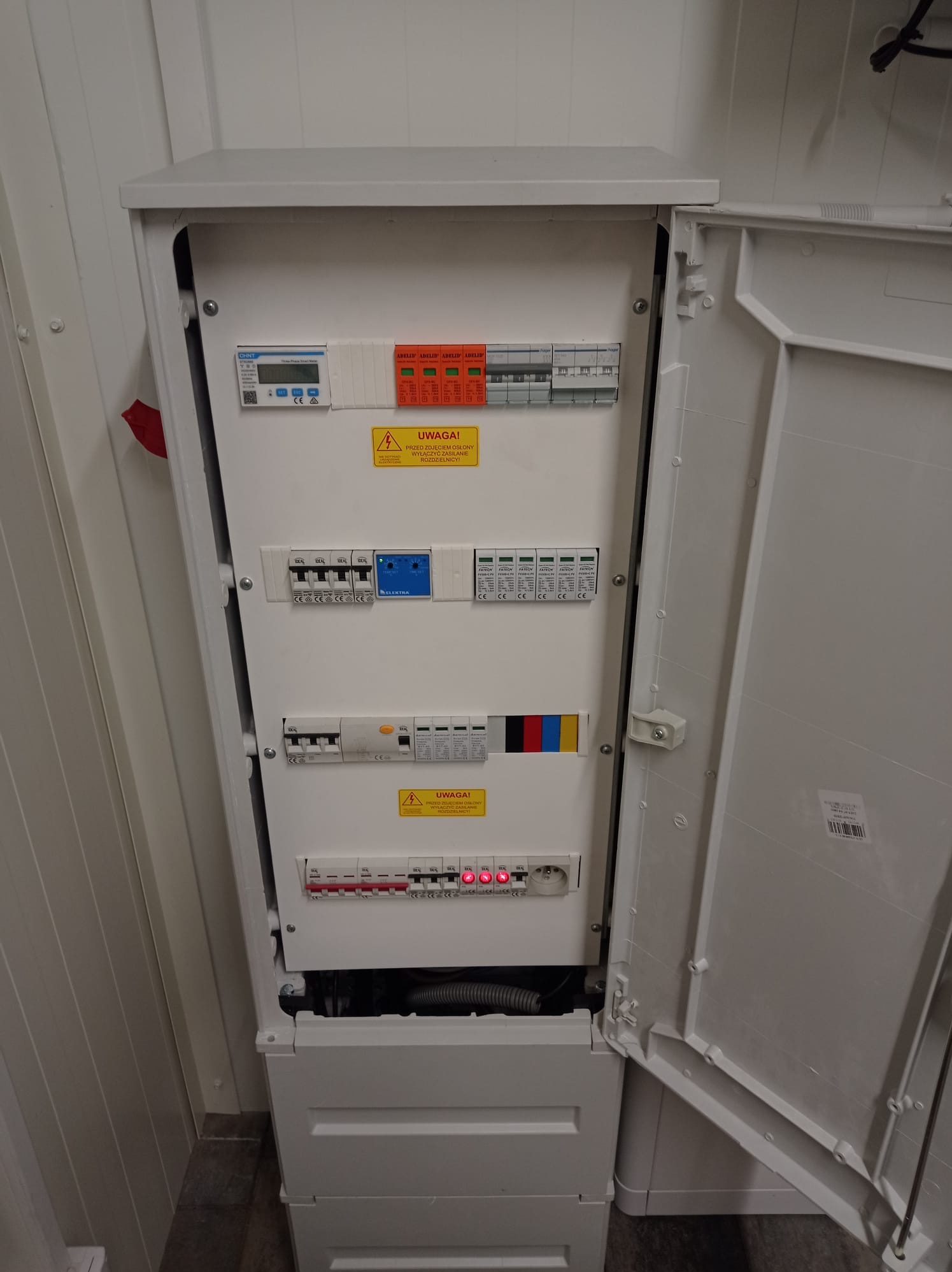 Otwarta, biała skrzynka rozdzielcza z widocznymi zabezpieczeniami, licznikiem i ostrzeżeniami. Widoczne kable i elementy instalacji elektrycznej wewnątrz.