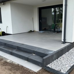 NETCORE CONNECT SPÓŁKA Z OGRANICZONĄ ODPOWIEDZIALNOŚCIĄ - Wykończony taras z szarych płyt, z czarnymi obrzeżami i schodami. Biały budynek w tle, ozdobne kamienie wzdłuż ściany. Widok na ogród.