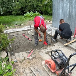 NETCORE CONNECT SPÓŁKA Z OGRANICZONĄ ODPOWIEDZIALNOŚCIĄ - Dwóch brukarzy w trakcie układania kostki. Widoczny zagęszczarka, poziomica i narzędzia. Częściowo ułożona kostka, teren przygotowany pod dalsze prace.