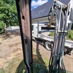 NETCORE CONNECT SPÓŁKA Z OGRANICZONĄ ODPOWIEDZIALNOŚCIĄ - Widok z kabiny koparki na wywrotkę z ziemią na tle domu z szarą dachówką i zielenią. Prace ziemne w toku, widoczny operator i teren.