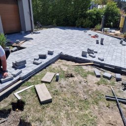 NETCORE CONNECT SPÓŁKA Z OGRANICZONĄ ODPOWIEDZIALNOŚCIĄ - Fragment układania szarej kostki brukowej na podjeździe domu jednorodzinnego. Widoczne narzędzia i częściowo ułożona nawierzchnia. Prace w toku, dzień słoneczny.