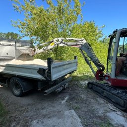 NETCORE CONNECT SPÓŁKA Z OGRANICZONĄ ODPOWIEDZIALNOŚCIĄ - Mini koparka ładuje piasek do skrzyni samochodu ciężarowego na tle zieleni. Operator koparki w kabinie. Praca w toku, słoneczny dzień.