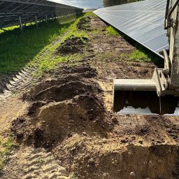 Kujawa Krystian NETCORE CONNECT - Wykop pod panele słoneczne wykonany koparką na terenie farmy fotowoltaicznej. Ślady opon na ziemi, pochmurne niebo w tle. Prace ziemne w toku.