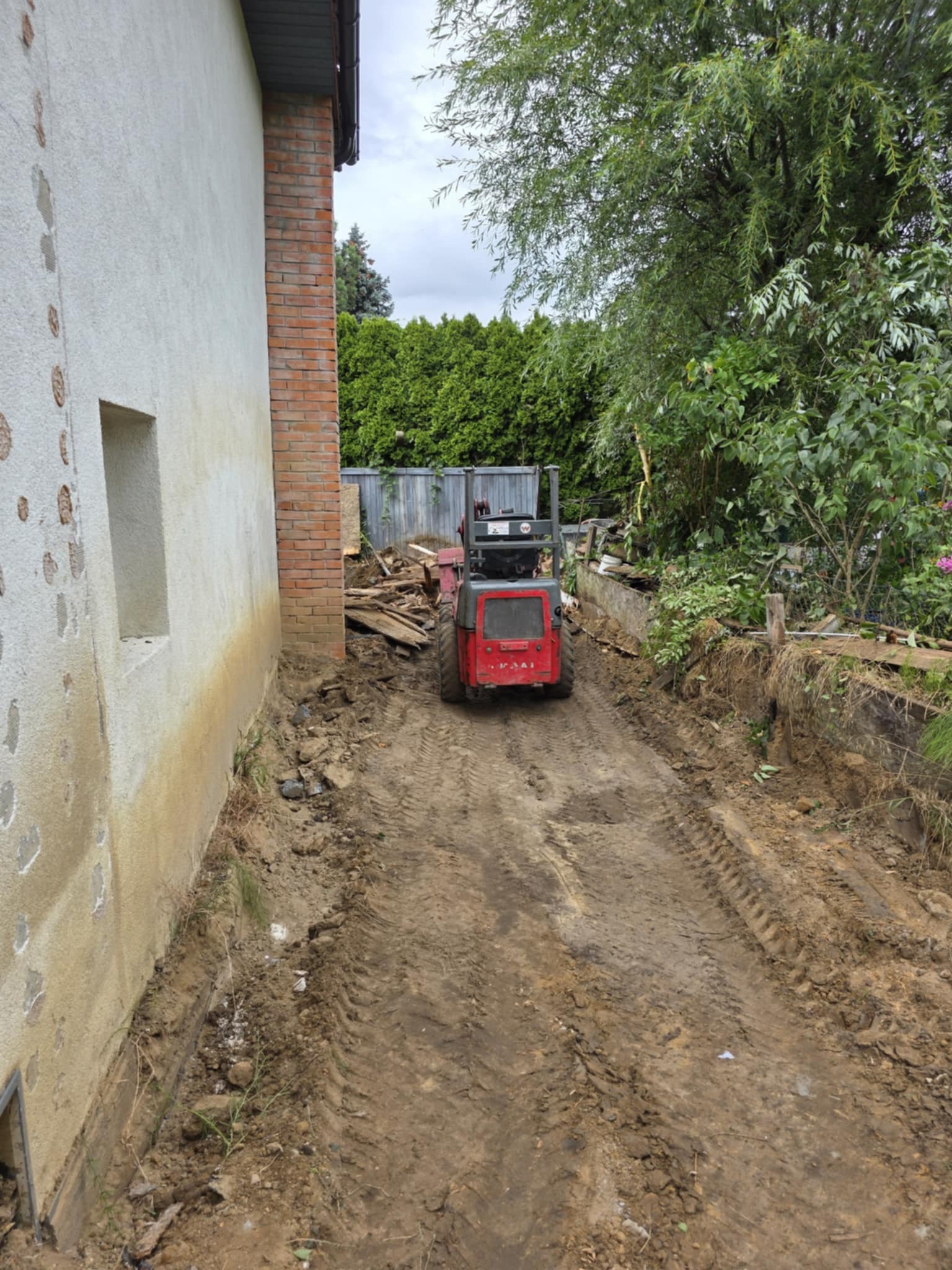 Czerwony mini-traktor na błotnistej drodze obok budynku z uszkodzonym tynkiem. Ślady opon w ziemi, gruz i roślinność w tle. Prace ziemne w toku.