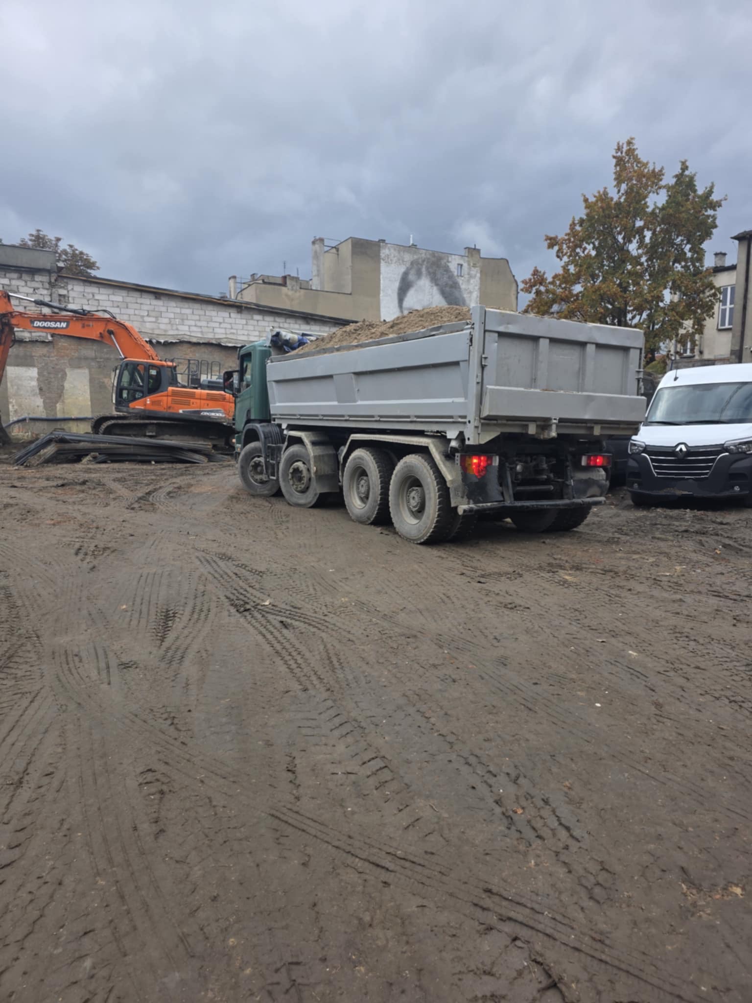 Ciężarówka z piaskiem i pomarańczowa koparka Doosan na placu budowy z ubitym, błotnistym podłożem. W tle budynki i biały samochód dostawczy.