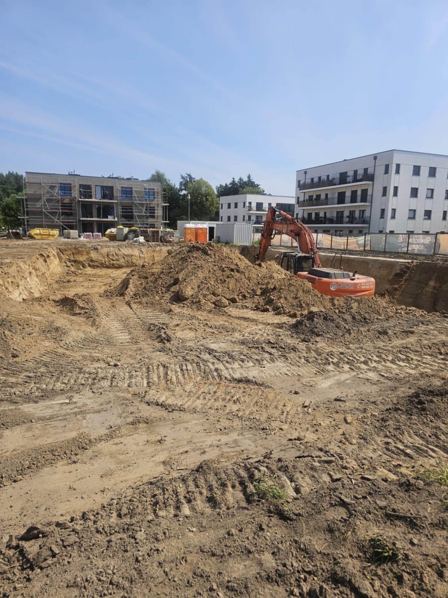 Pomarańczowa koparka URUS w wykopie na placu budowy, w tle budynki w stanie surowym i kontenery. Ślady opon na ziemi, błękitne niebo.