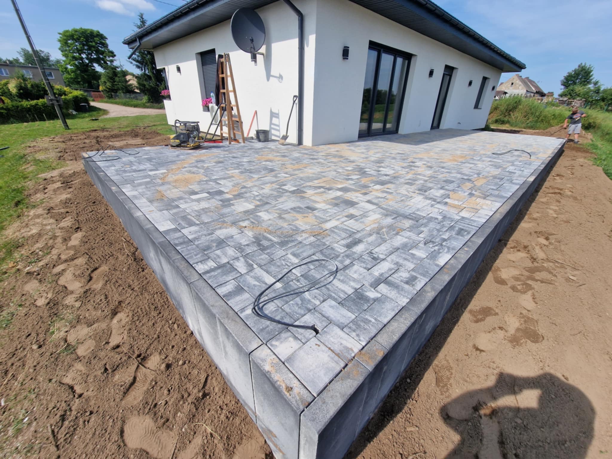 Nowoczesny taras z szarej kostki brukowej ułożonej w geometryczny wzór, z widocznym obrzeżem i przygotowanym terenem pod dalsze prace wykończeniowe.