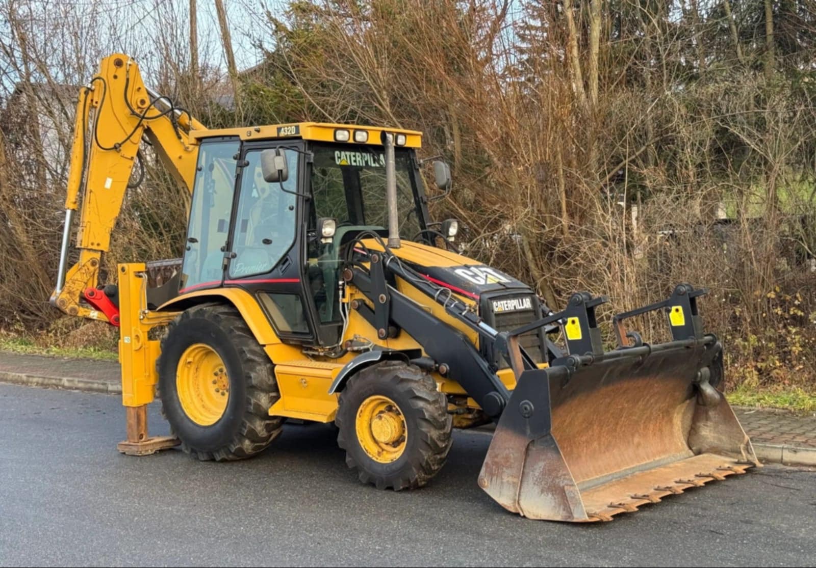 Żółto-czarna koparko-ładowarka Caterpillar 432D na asfaltowej drodze, z podniesionym ramieniem koparki i opuszczoną łyżką, na tle drzew bez liści.