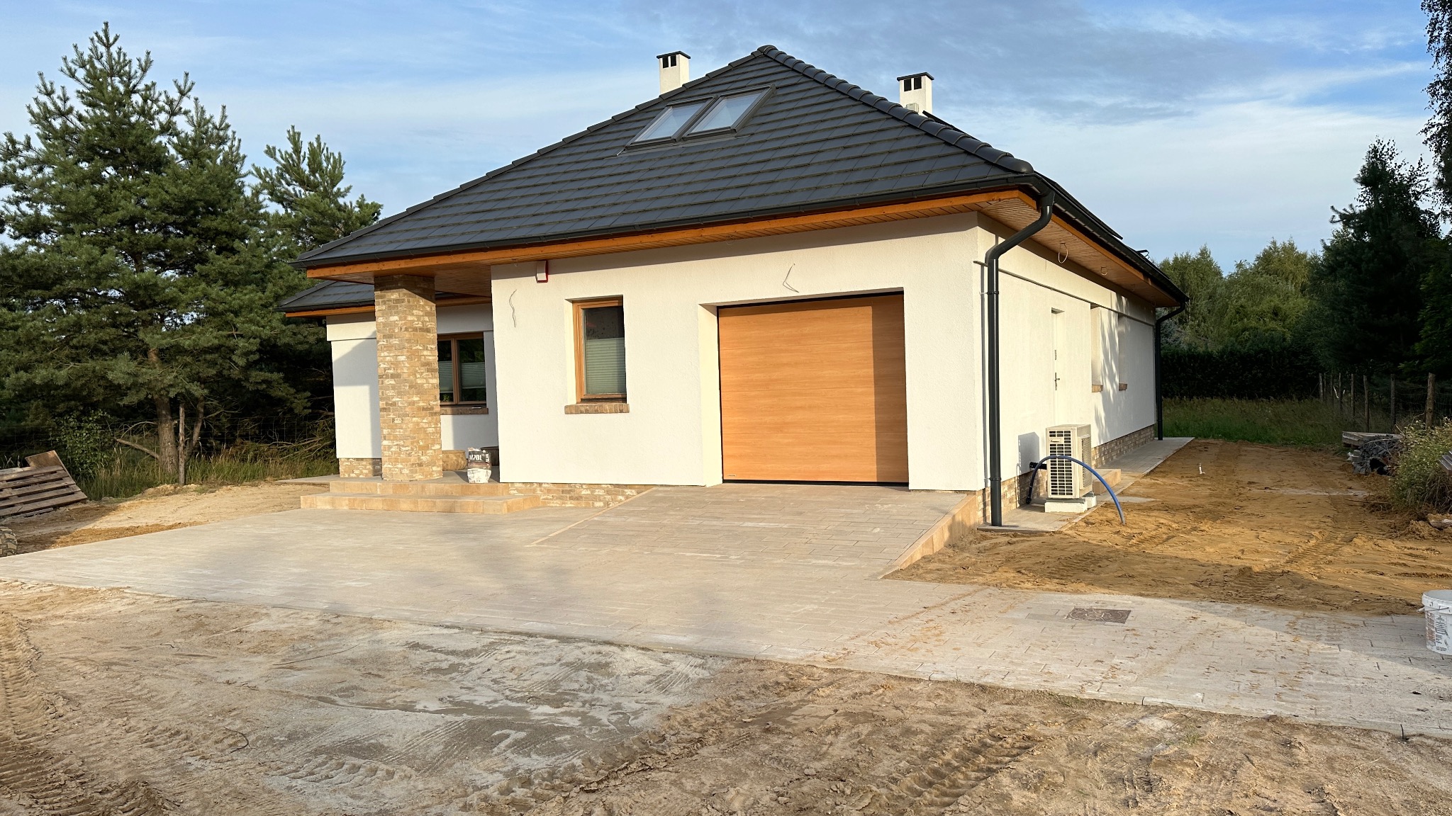 Nowoczesny dom jednorodzinny z garażem i podjazdem z kostki brukowej w trakcie wykańczania. Widoczne elementy elewacji i otoczenie w stanie surowym.
