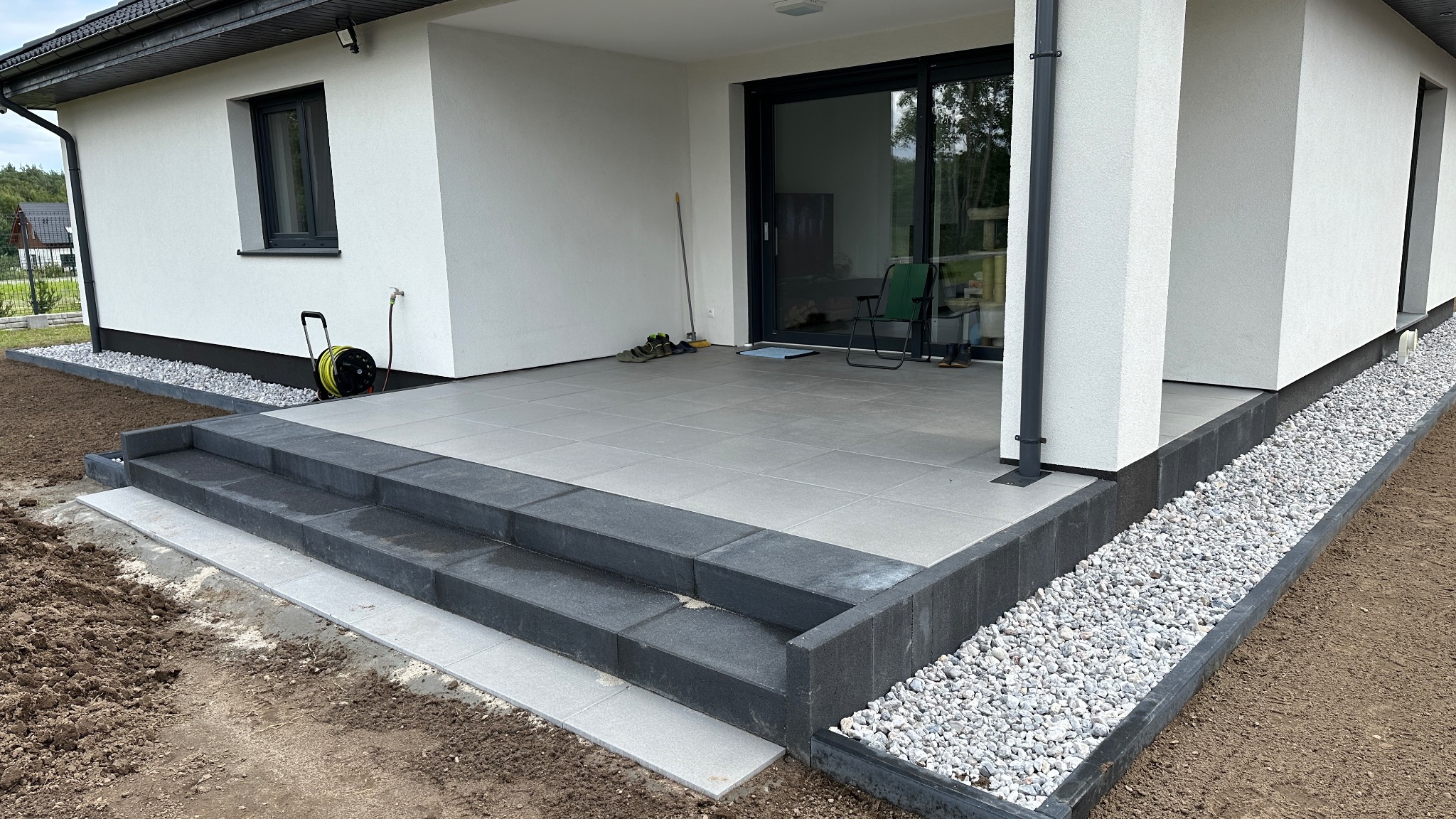 Wykończony taras z szarych płyt, z czarnymi obrzeżami i schodami. Biały budynek w tle, ozdobne kamienie wzdłuż ściany. Widok na ogród.