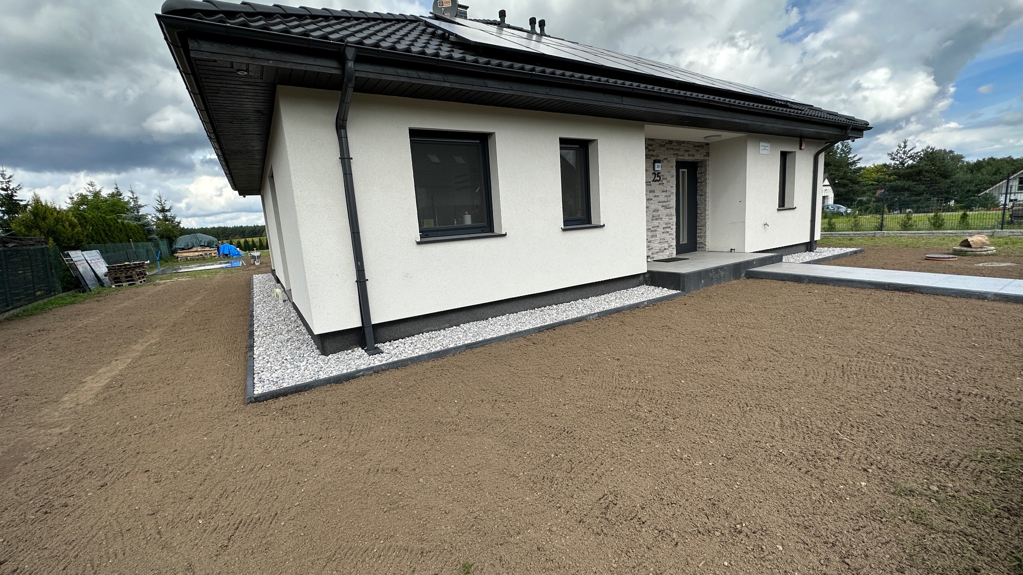 Nowoczesny dom jednorodzinny z białym tynkiem, czarną stolarką i dachem z panelami słonecznymi. Wokół domu wysypany biały kamień, przygotowany teren pod trawnik.