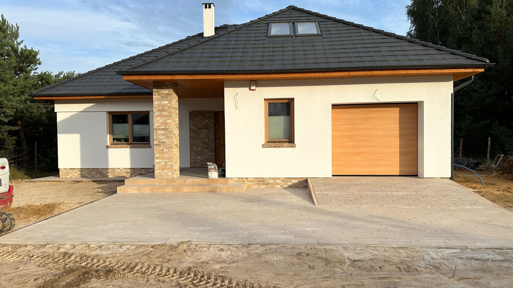 Nowoczesny dom jednorodzinny z jasną elewacją i brukowanym podjazdem. Widoczne detale wykończenia, takie jak drewniana brama garażowa i ceglana kolumna.