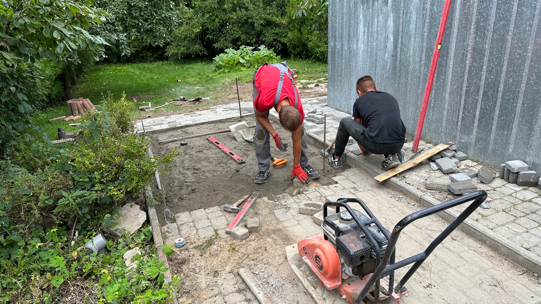 Dwóch brukarzy w trakcie układania kostki. Widoczny zagęszczarka, poziomica i narzędzia. Częściowo ułożona kostka, teren przygotowany pod dalsze prace.