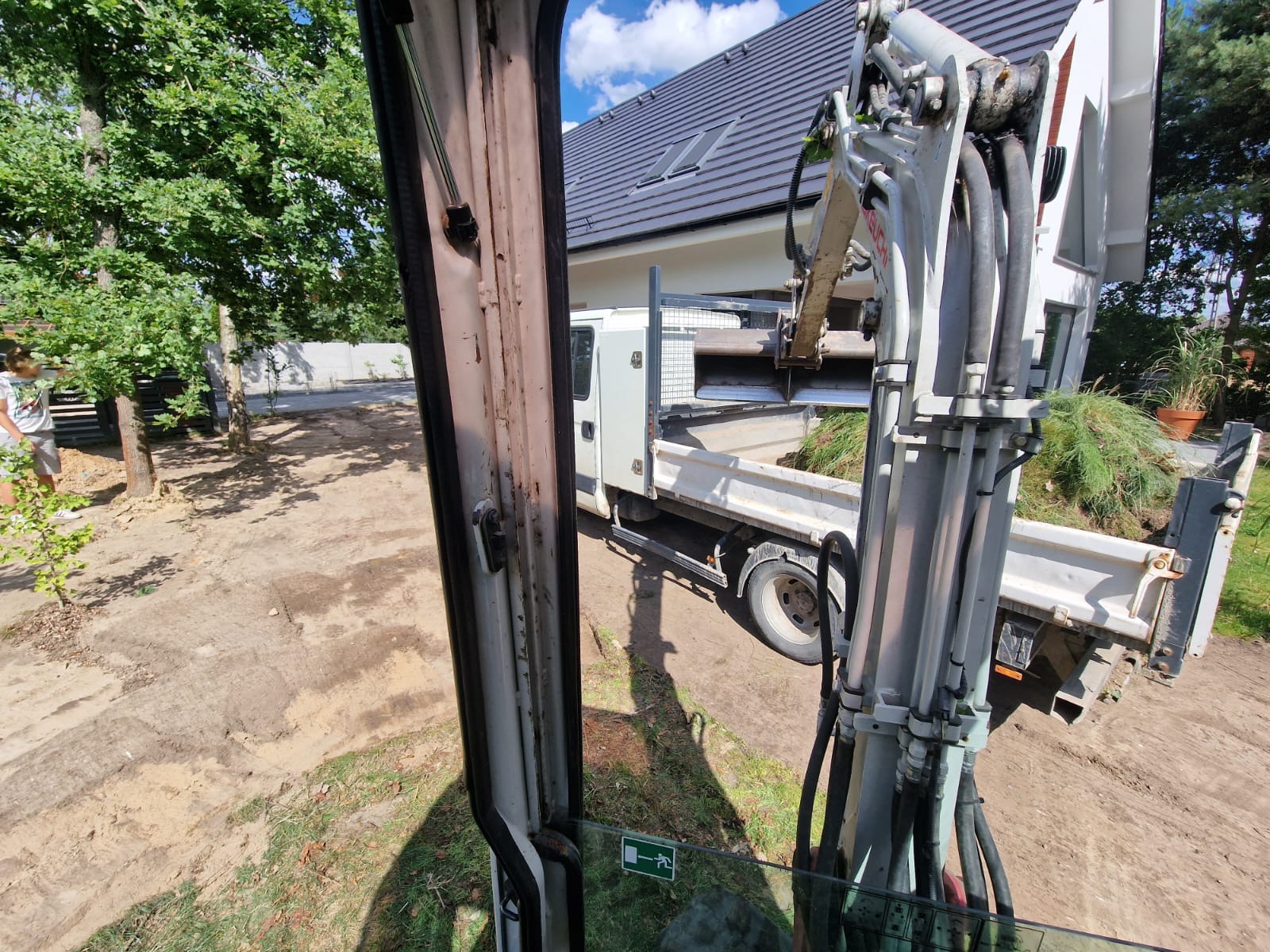 Widok z kabiny koparki na wywrotkę z ziemią na tle domu z szarą dachówką i zielenią. Prace ziemne w toku, widoczny operator i teren.
